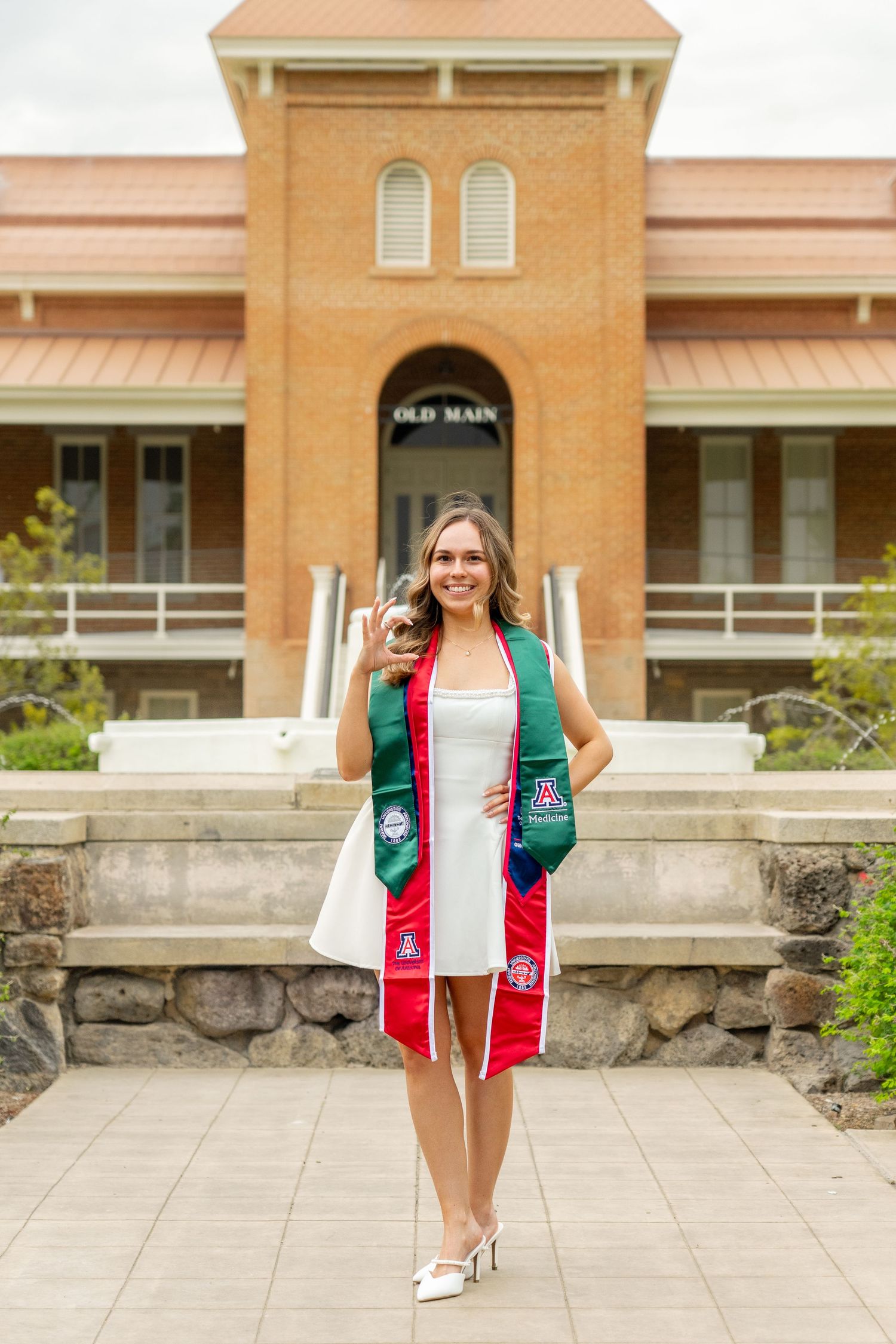 Smiling graduate in white dress with red and green stole poses in front of Old Main building on college campus.