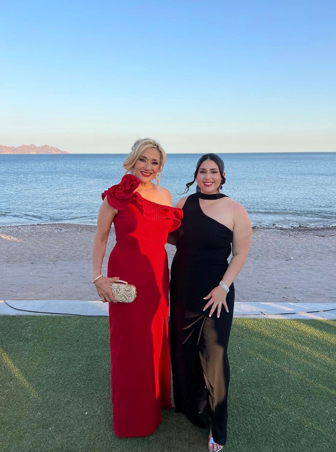 Beachside formal event with two people in red and black dresses against ocean backdrop.