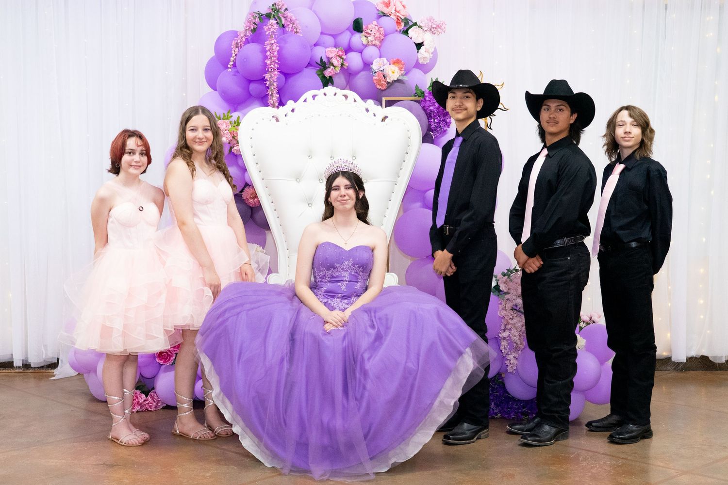 Quinceañera party posed in purple gowns and black suits against white backdrop.