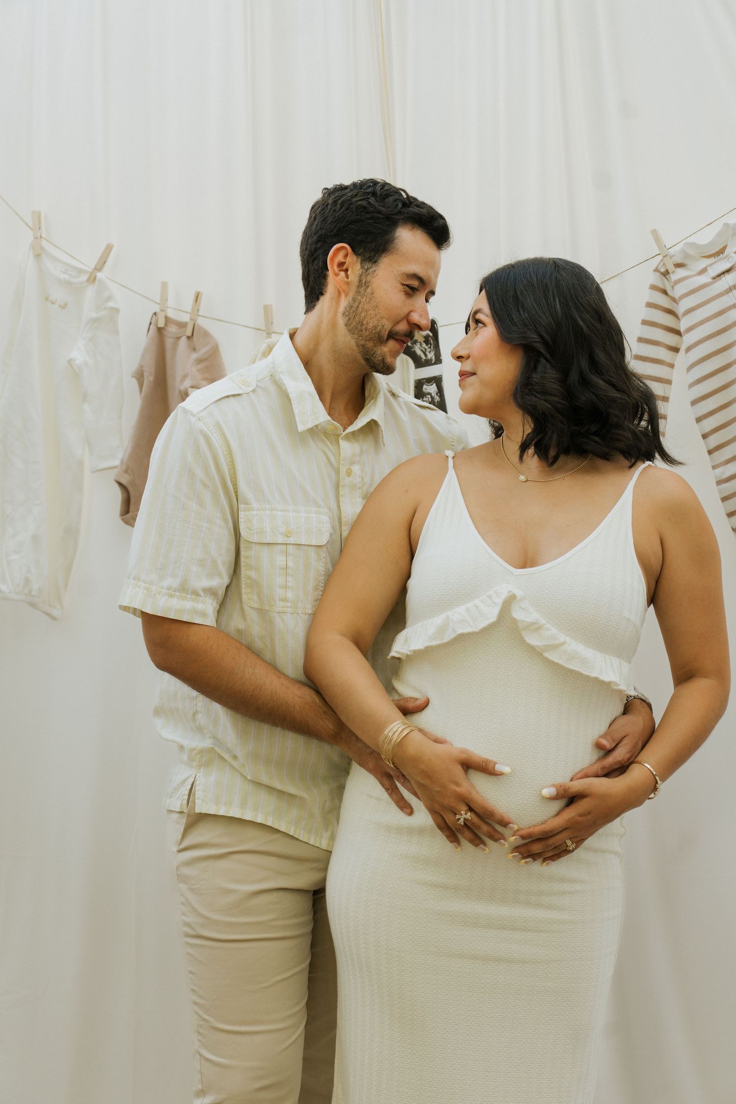 Couple in light colored outfits sharing tender moment during pregnancy photoshoot.