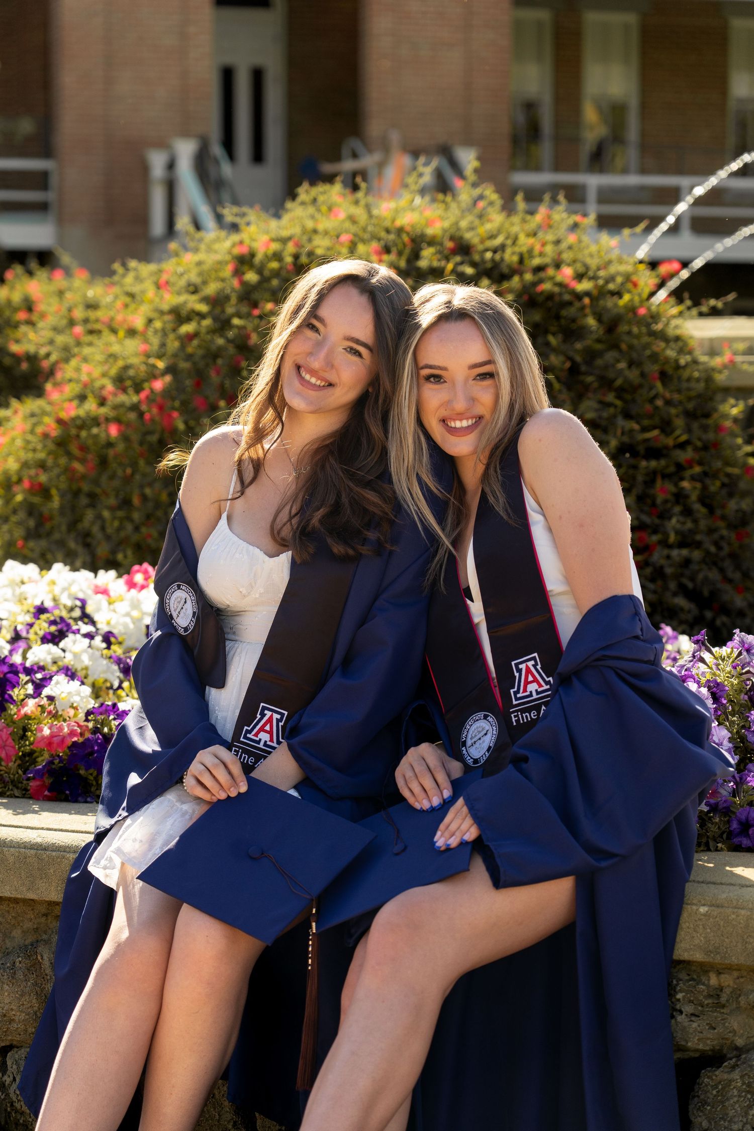 Sisters in graduation attire pose playfully near blooming flowers at Old Main on University of Arizona campus.