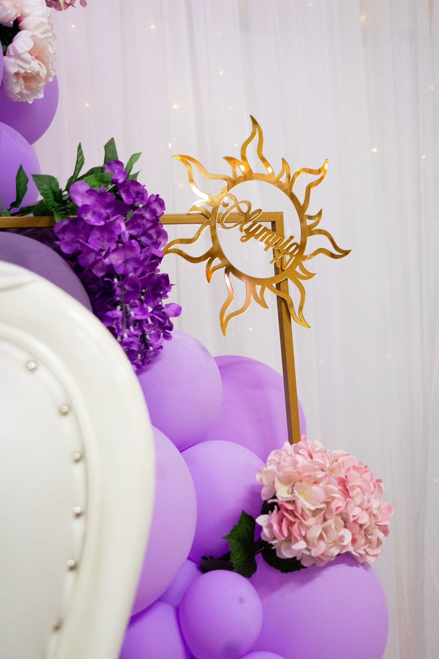 Purple balloons and gold sun decorations against a white backdrop with floral accents.