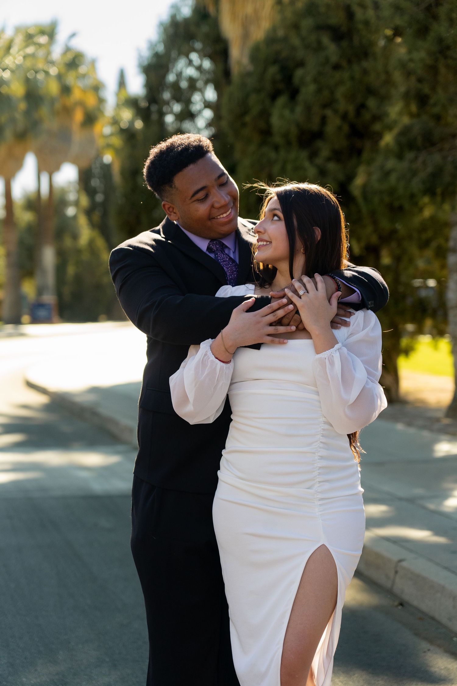 Couple embracing outdoors in formal attire with palm trees in background.
