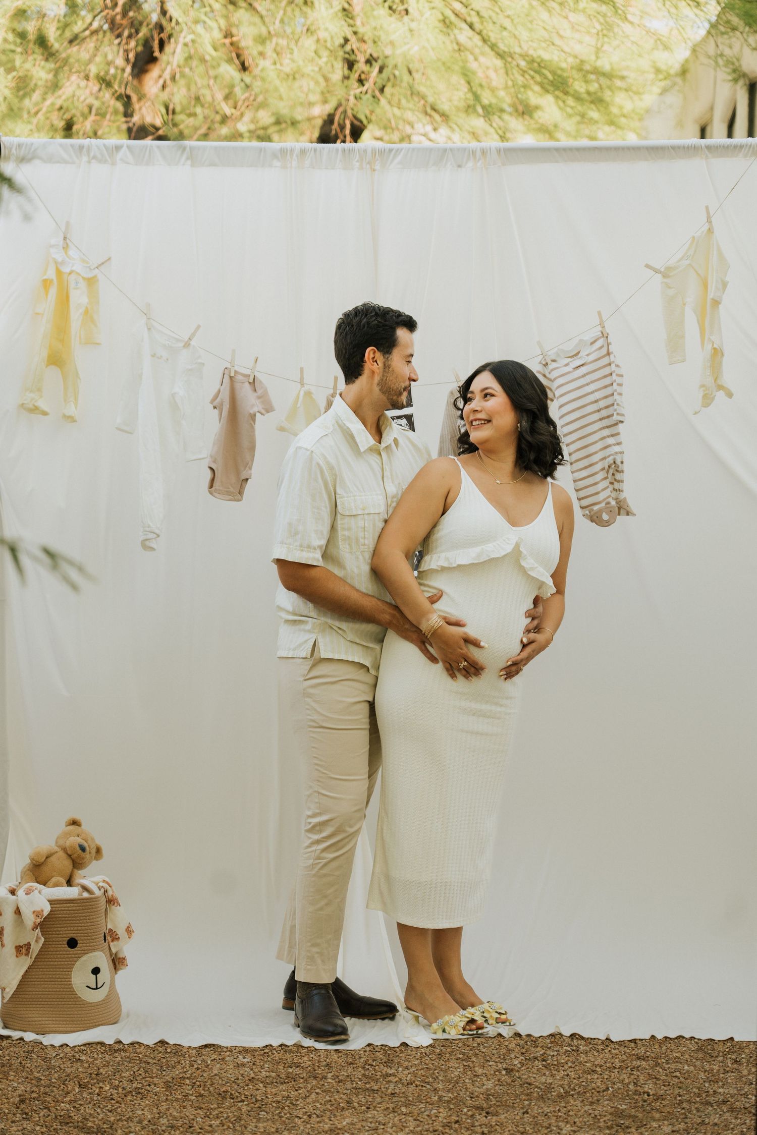 Expecting couple poses against white backdrop with baby clothes and teddy bear decorations.