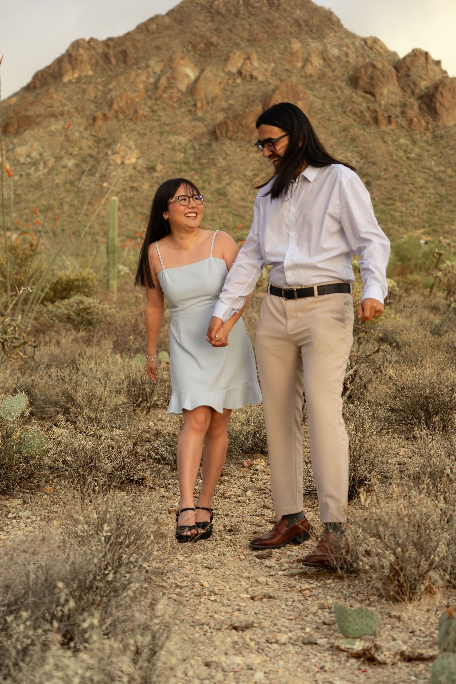 Couple walking and laughing together on desert trail with mountain and cactus landscape.