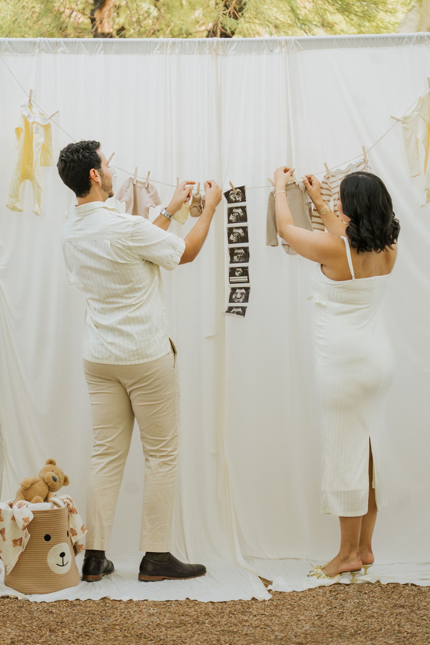 Couple hanging baby clothes and ultrasound photos on white backdrop outdoors.
