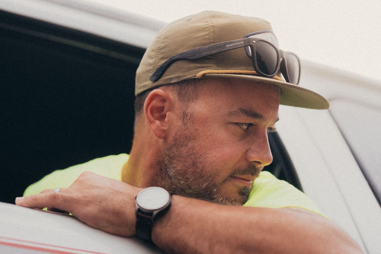 Man wearing a khaki cap, sunglasses, yellow shirt, and watch leaning on a white surface outdoors.