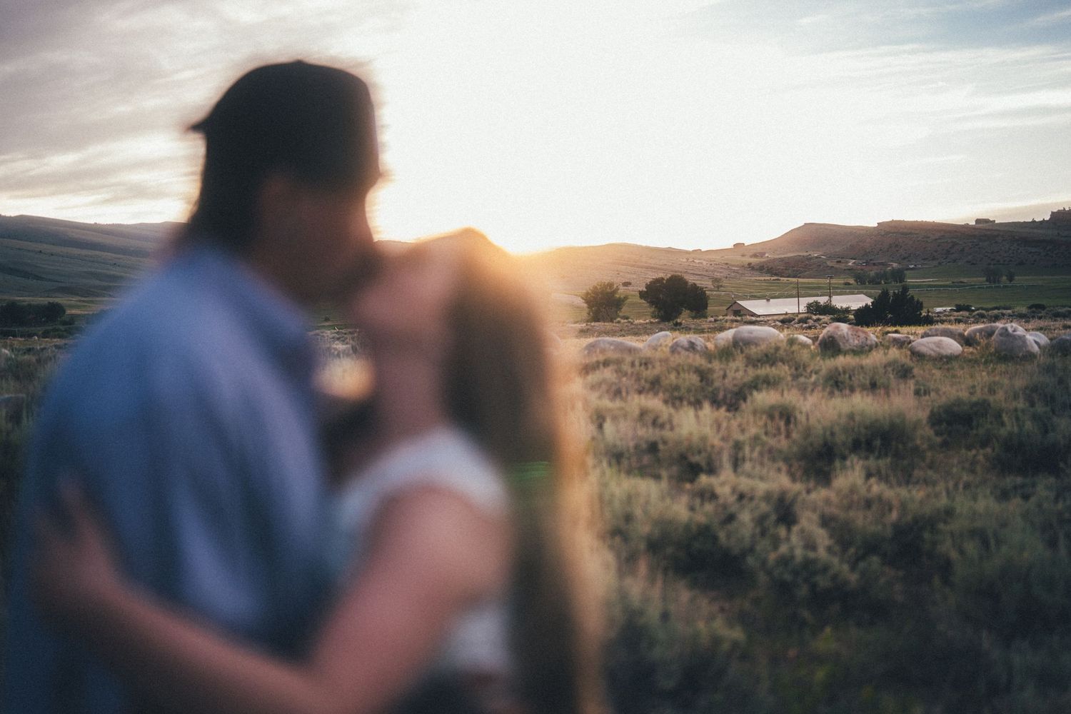 Couple sharing a romantic sunset kiss in a scenic rural landscape with golden light and rolling hills in the background.