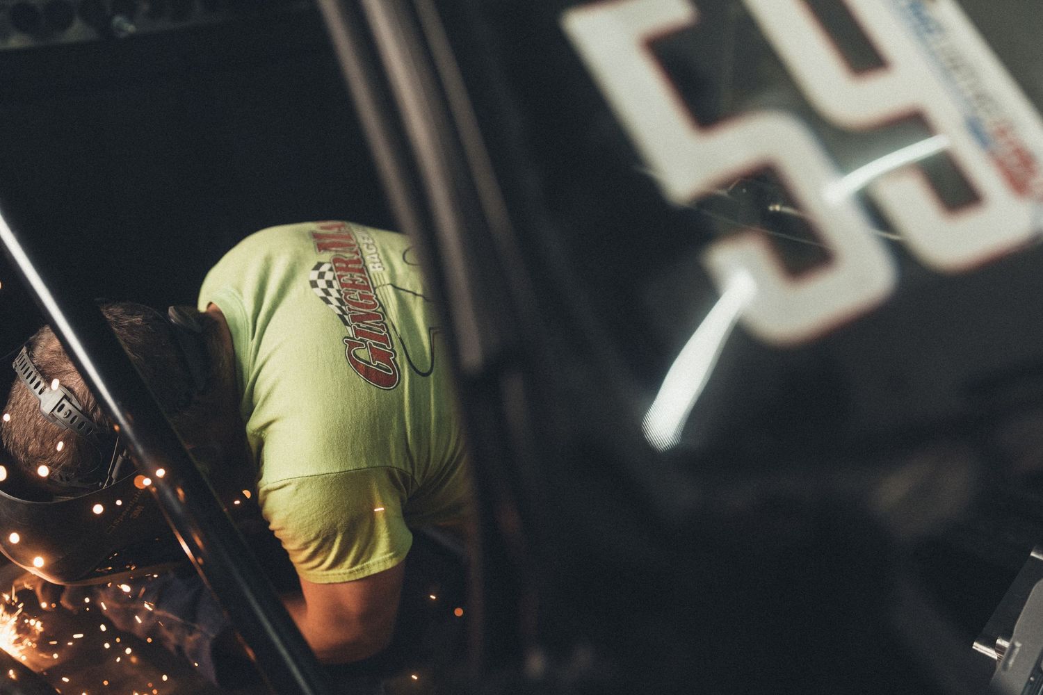 Sparks fly dramatically from a metal grinder while someone in a yellow t-shirt works in dark industrial setting.