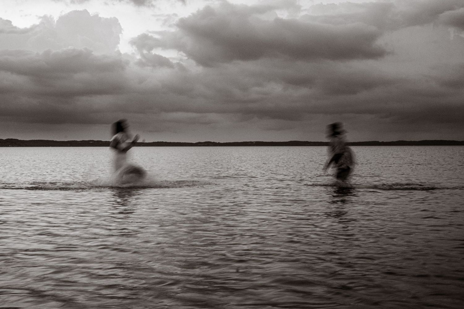 Motion-blurred figures move across a lake under dramatic cloudy skies in black and white.