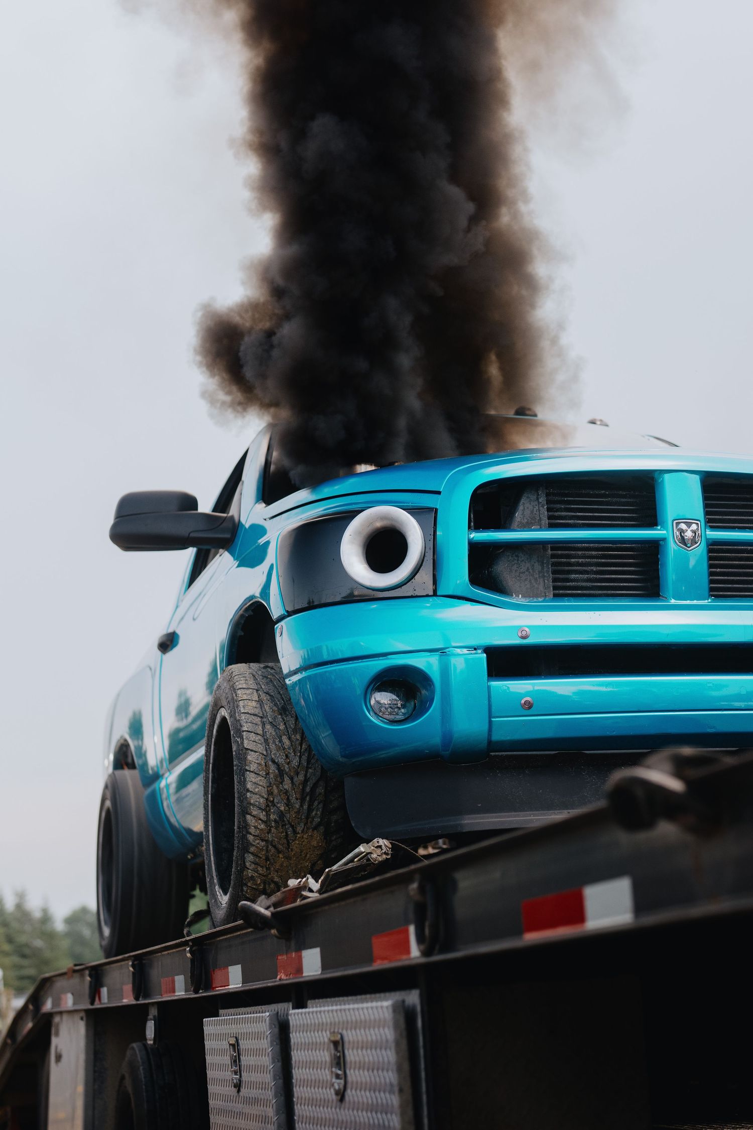 Blue Dodge Ram truck on flatbed tow truck emitting thick black smoke from engine fire.