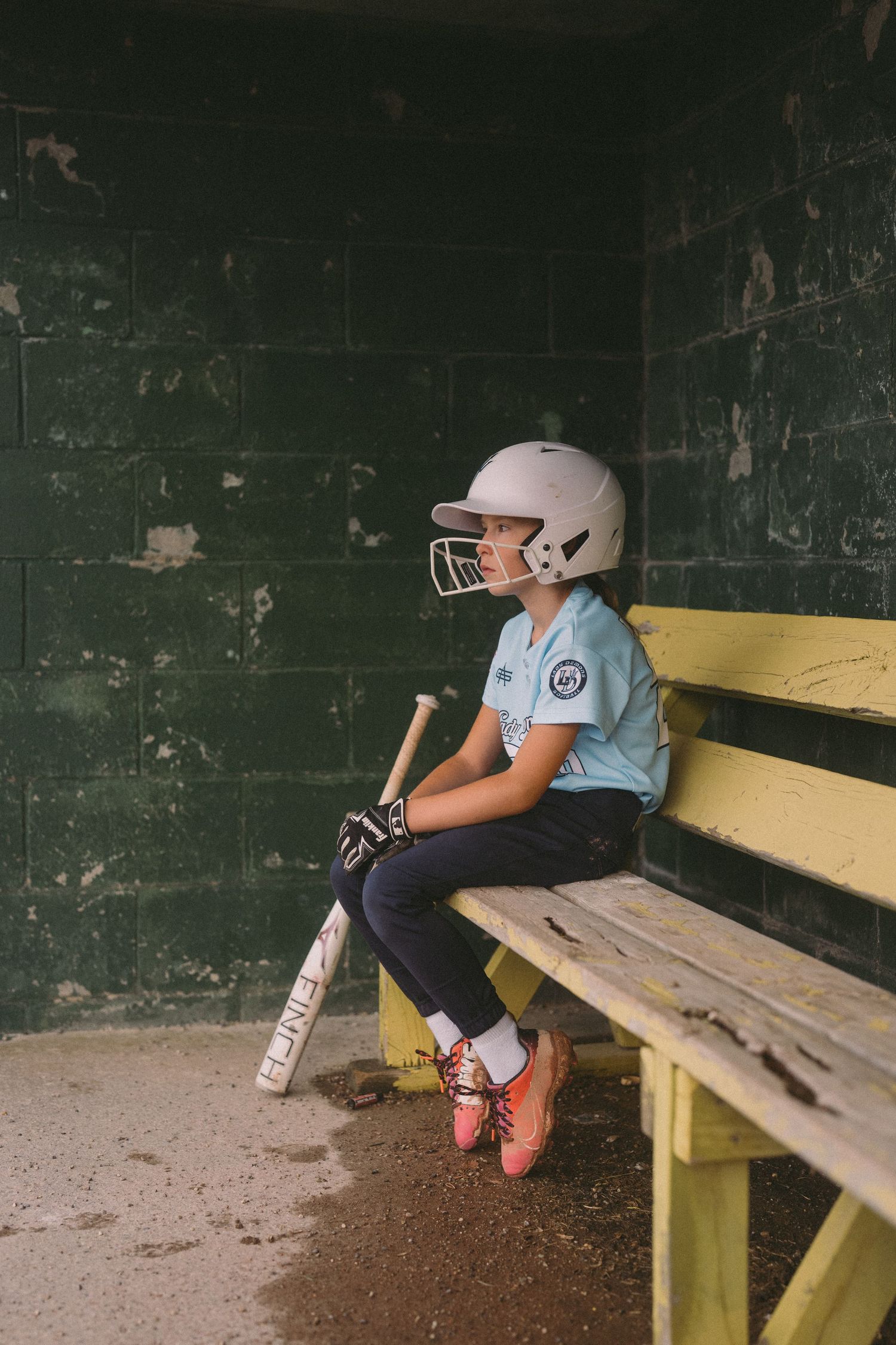 Young softball player in helmet sits in dugout holding bat, wearing light blue jersey and orange cleats.