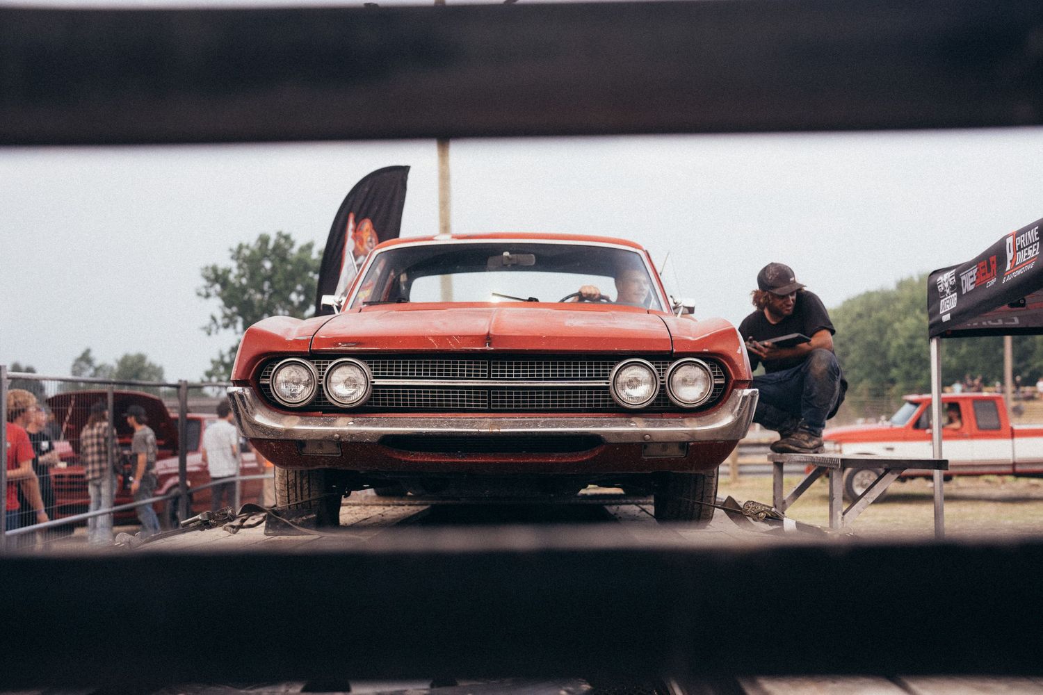 A vintage red muscle car on a drag strip with a mechanic leaning over the hood at an outdoor racing event.