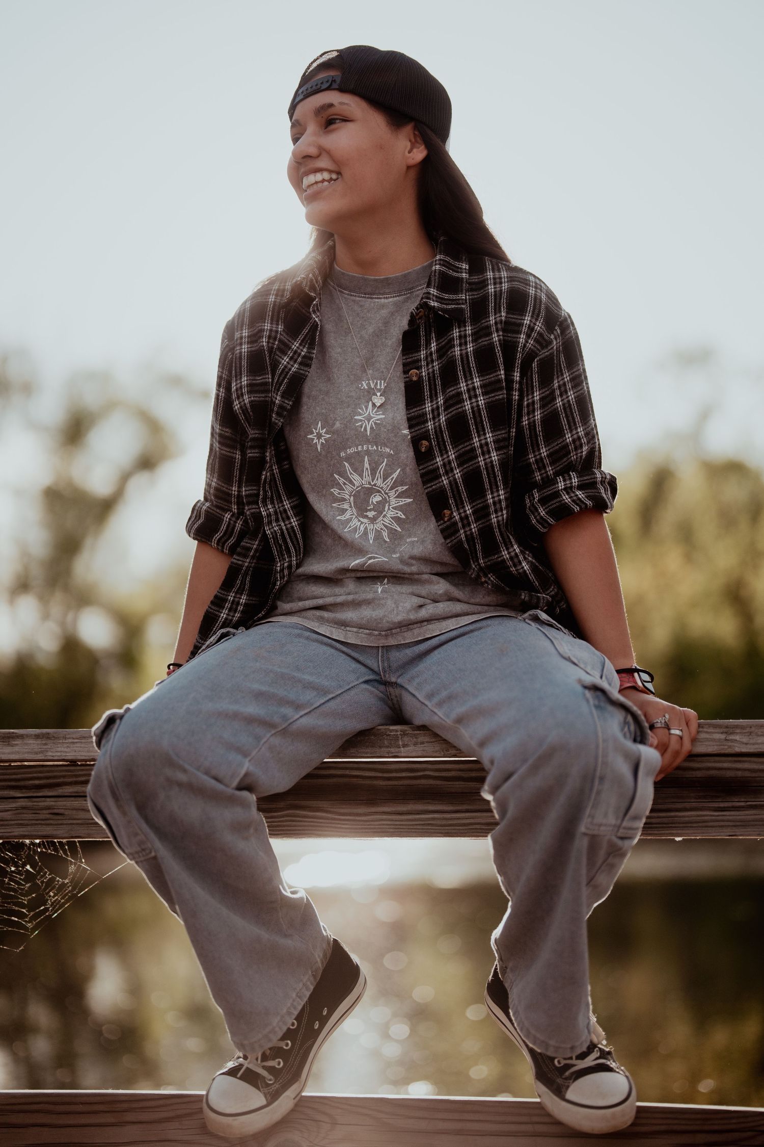 Person sitting casually on a wooden surface wearing plaid shirt and jeans at sunset.