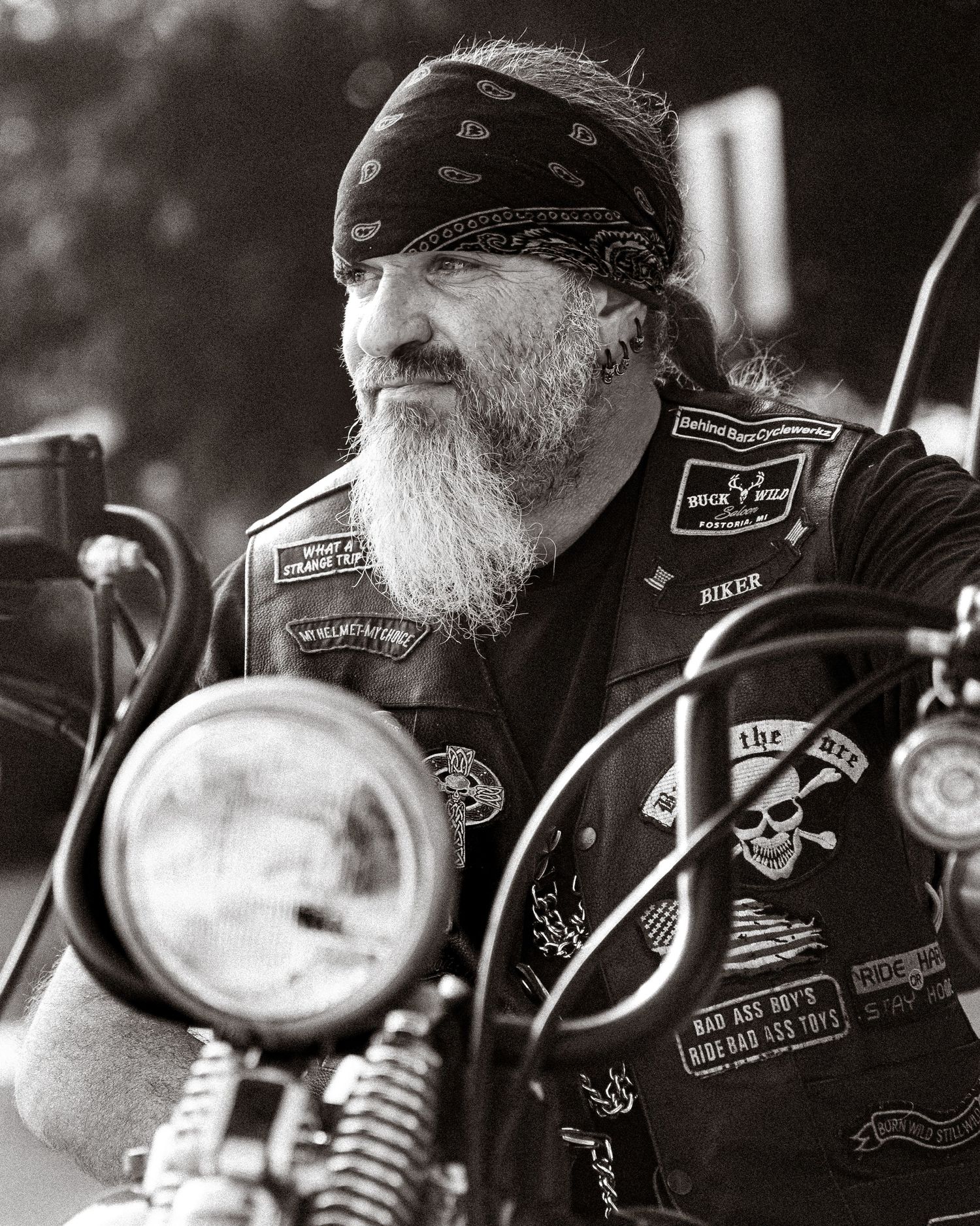 Black and white photo of biker with patches on leather vest sitting on motorcycle.