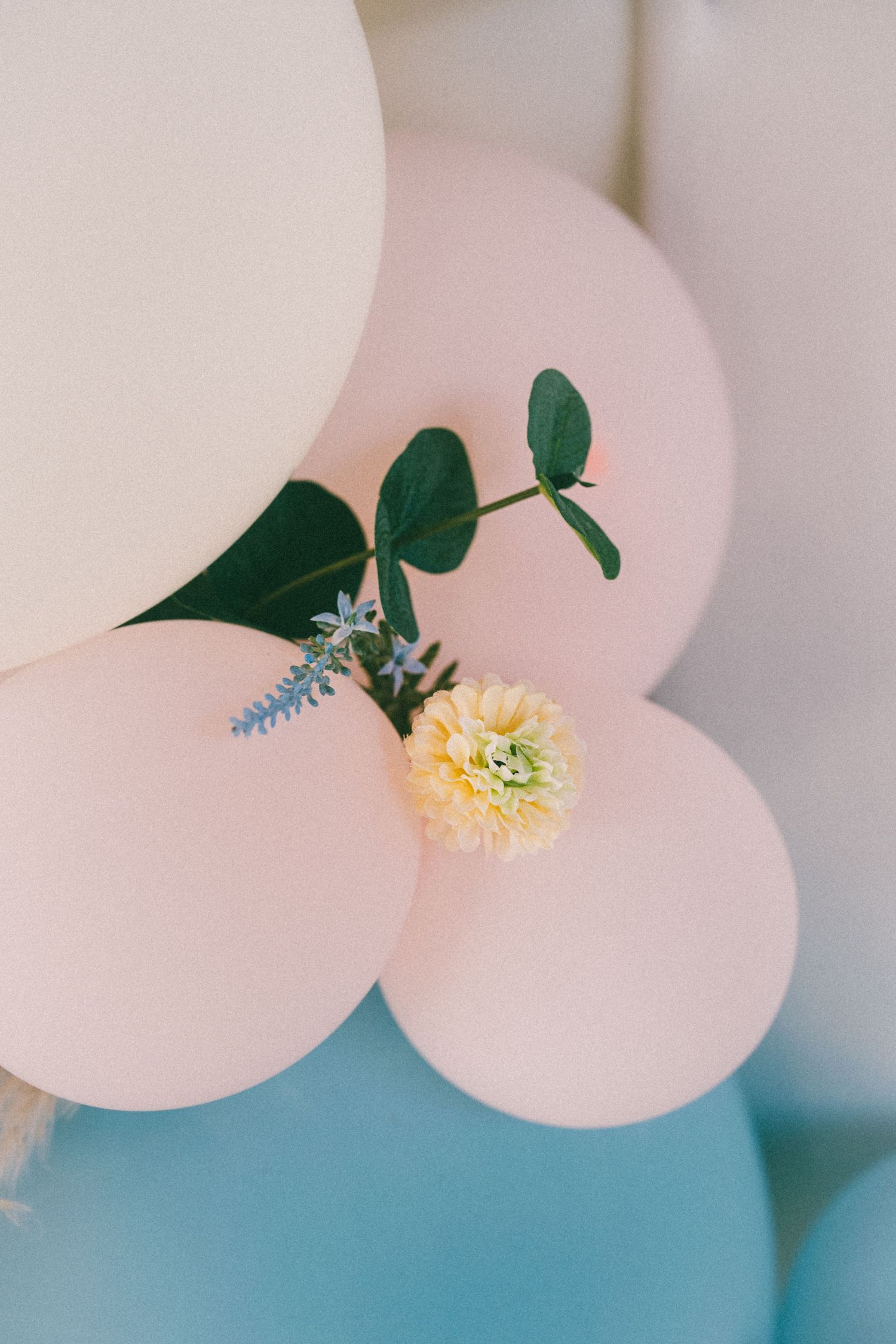 Pastel pink balloons decorated with a yellow dahlia, blue flowers, and green eucalyptus leaves against a light blue background.