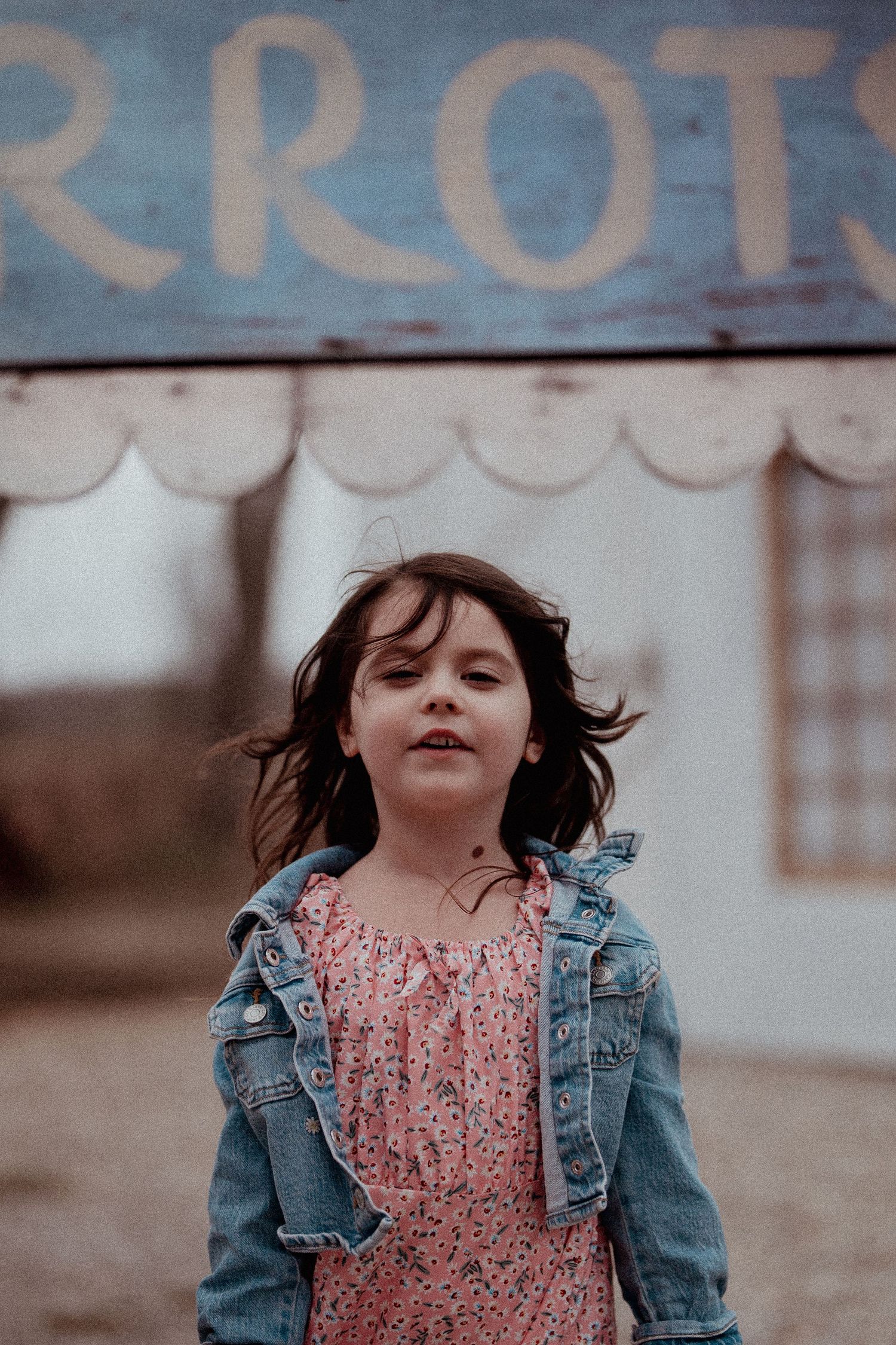 Girl in denim jacket and floral dress smiling joyfully outdoors.