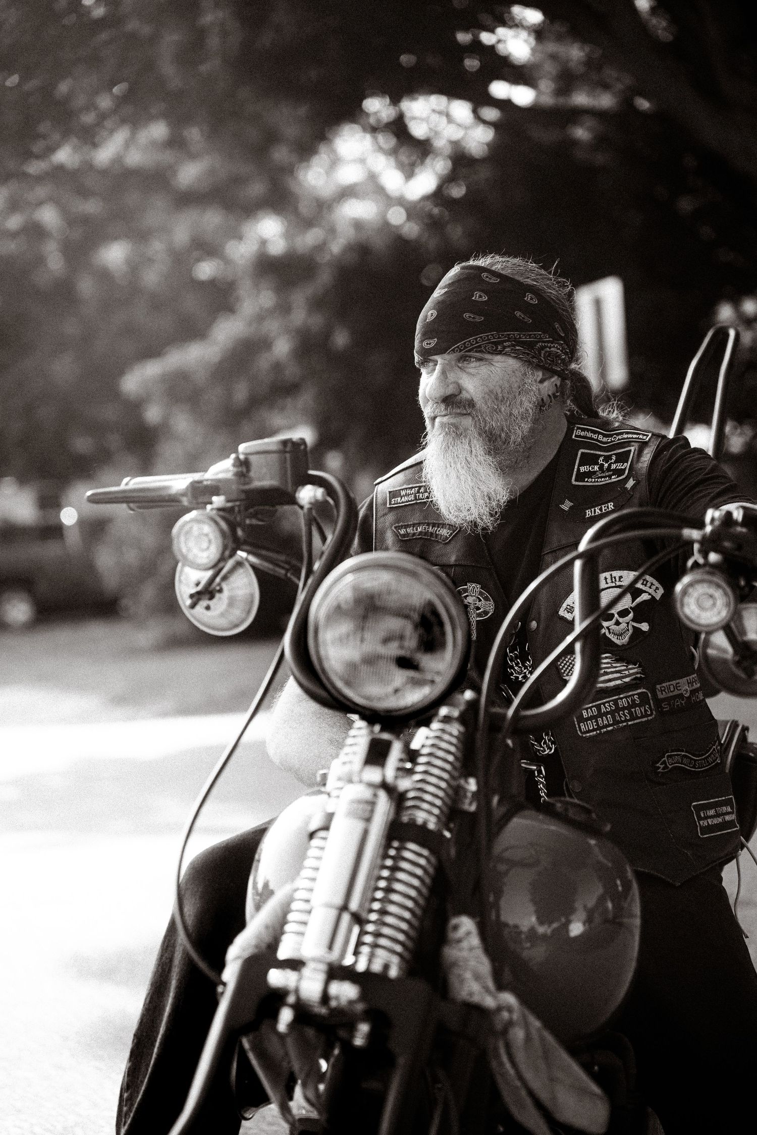 Black and white photo of a bearded biker with bandana sitting on a decorated motorcycle outdoors.