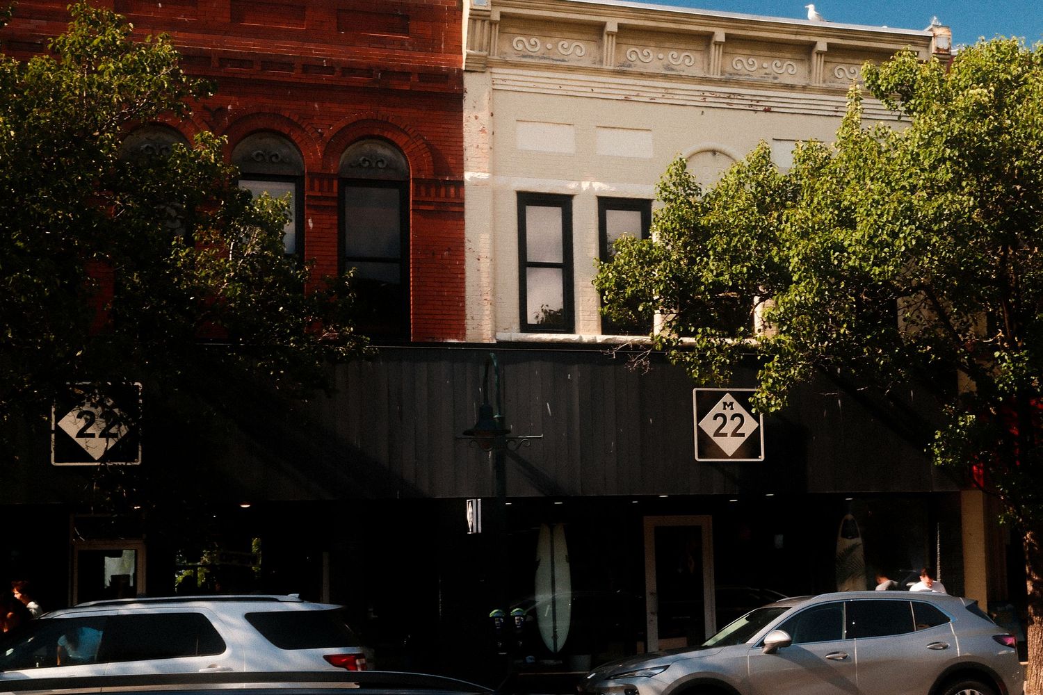 Storefront view of red brick buildings with black trim and trees along a city street.