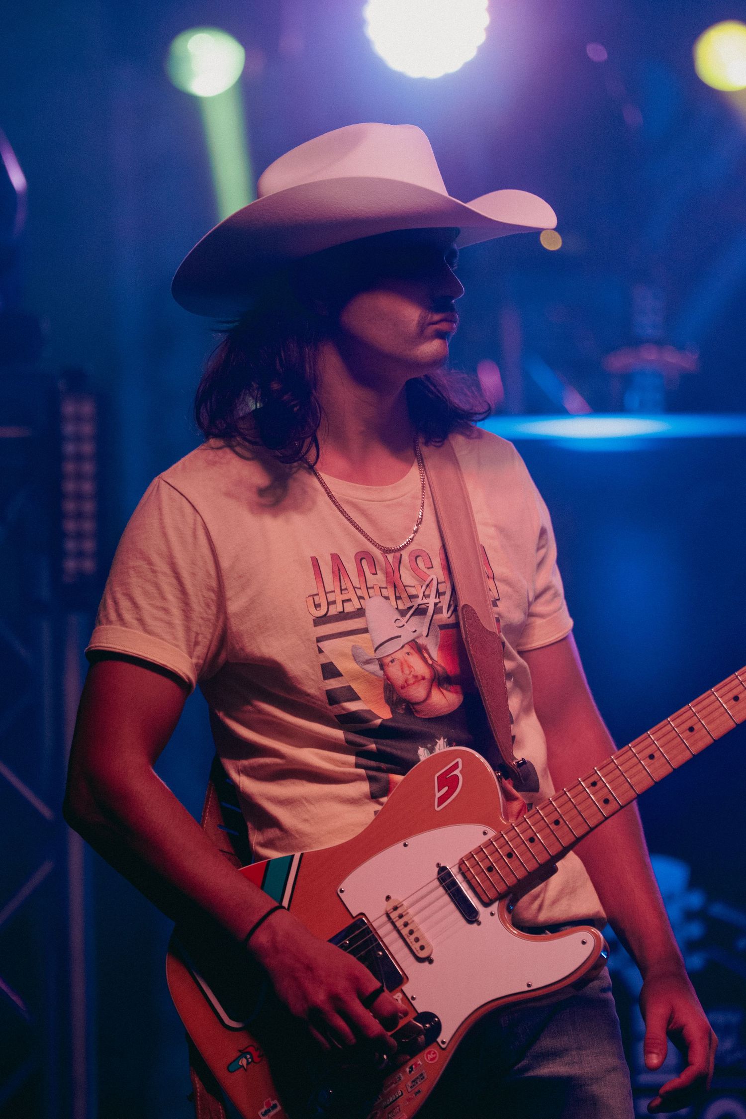 Guitar player wearing a cowboy hat performing on stage under blue concert lighting.