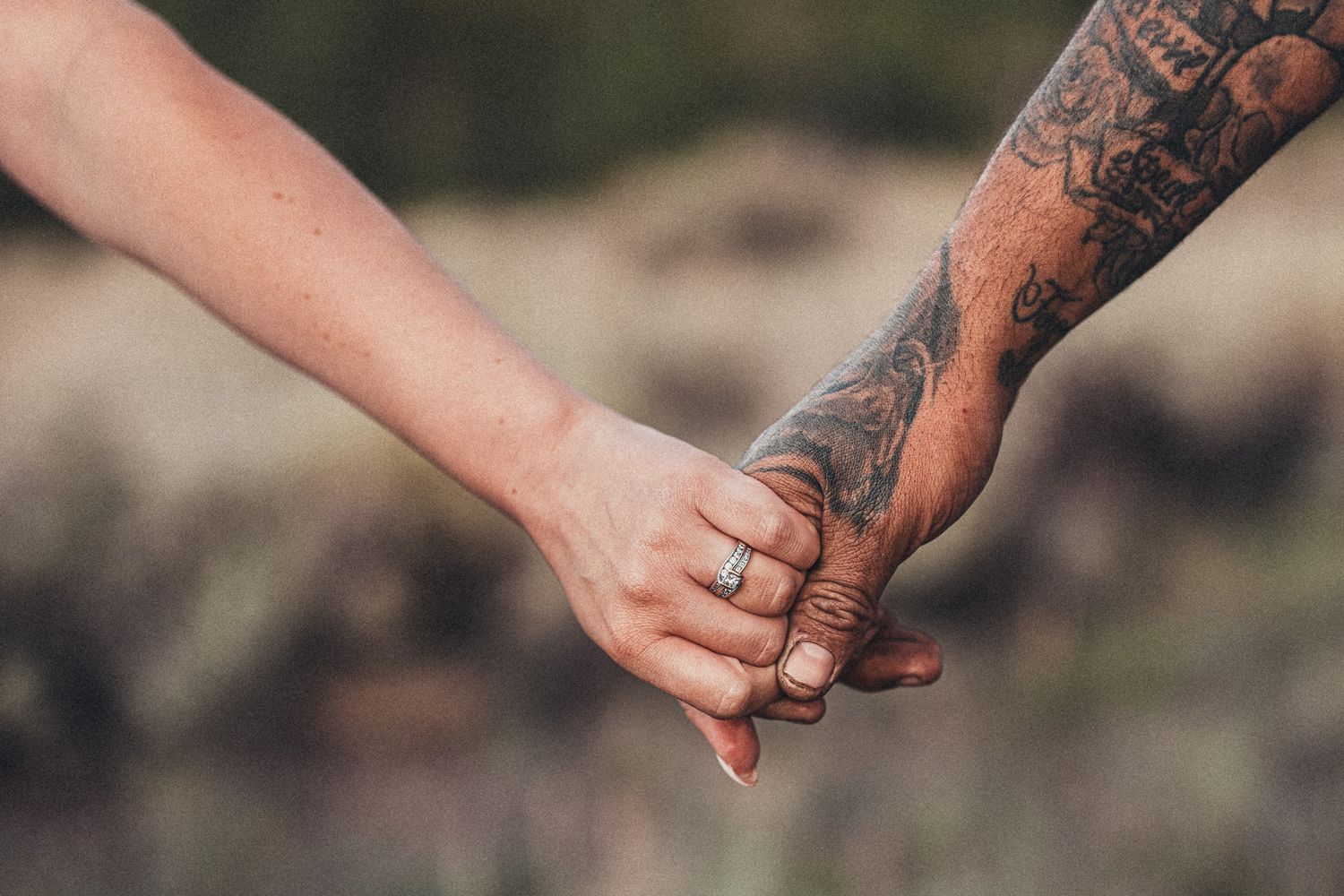 Close-up of a couple holding hands, woman wearing engagement ring, man with tattoo sleeve, romantic outdoor setting.