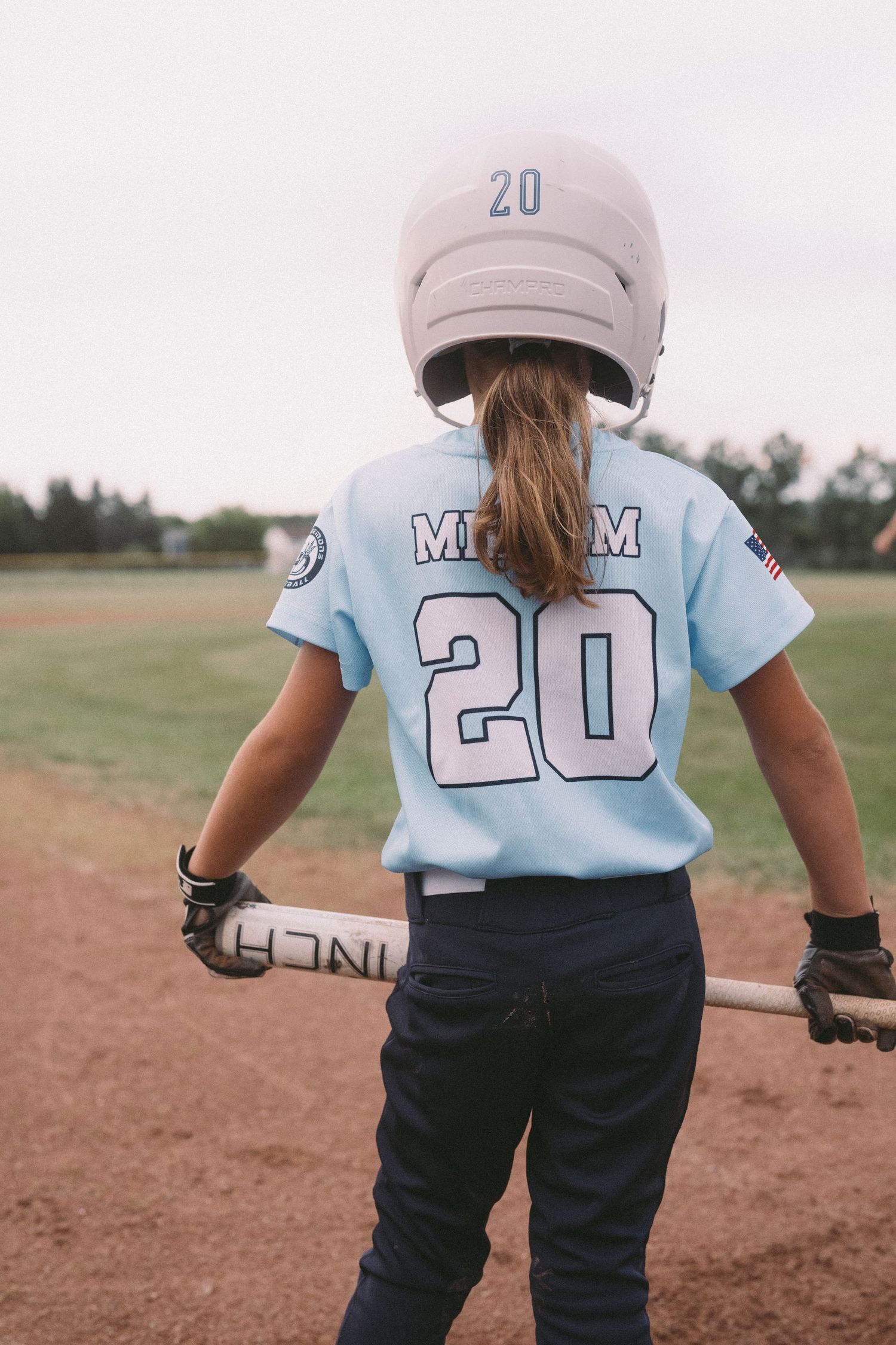 Young girl in light blue baseball jersey number 20 holding bat on field.