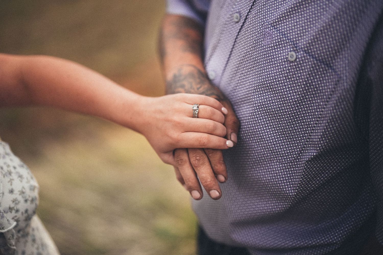 Close-up of a couple holding hands, woman wearing a diamond engagement ring, man with a tattoo on his forearm.