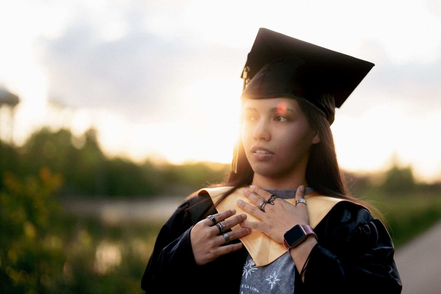Graduate in black cap and gown poses outdoors during golden hour sunset graduation photoshoot.