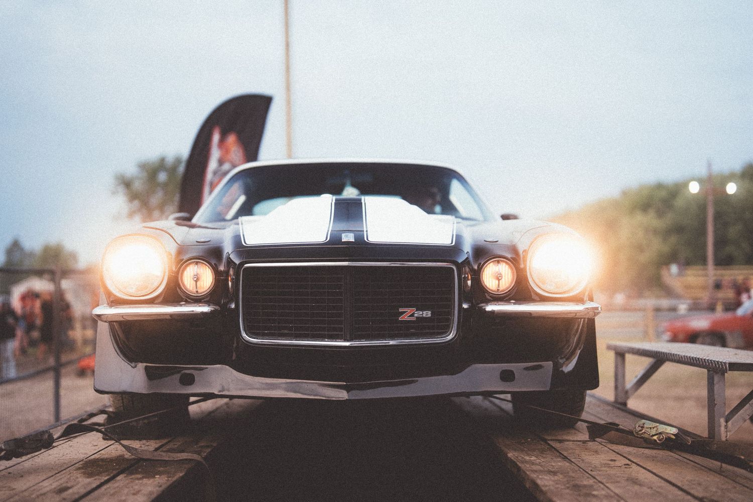 A classic Chevrolet Camaro Z28 muscle car with glowing headlights parked at an outdoor car show at dusk.