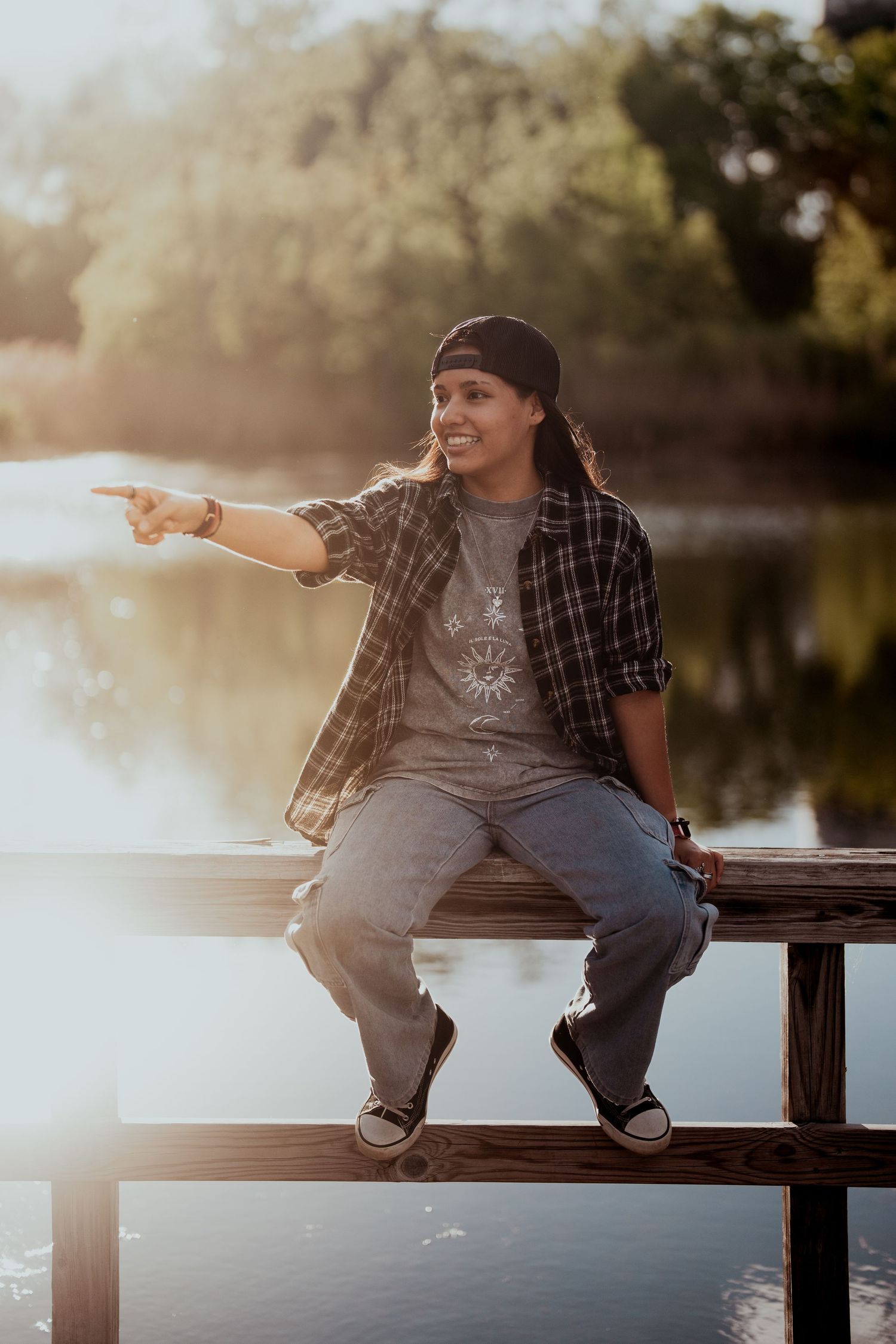 Person pointing while sitting on dock wearing plaid shirt and beanie at sunset.