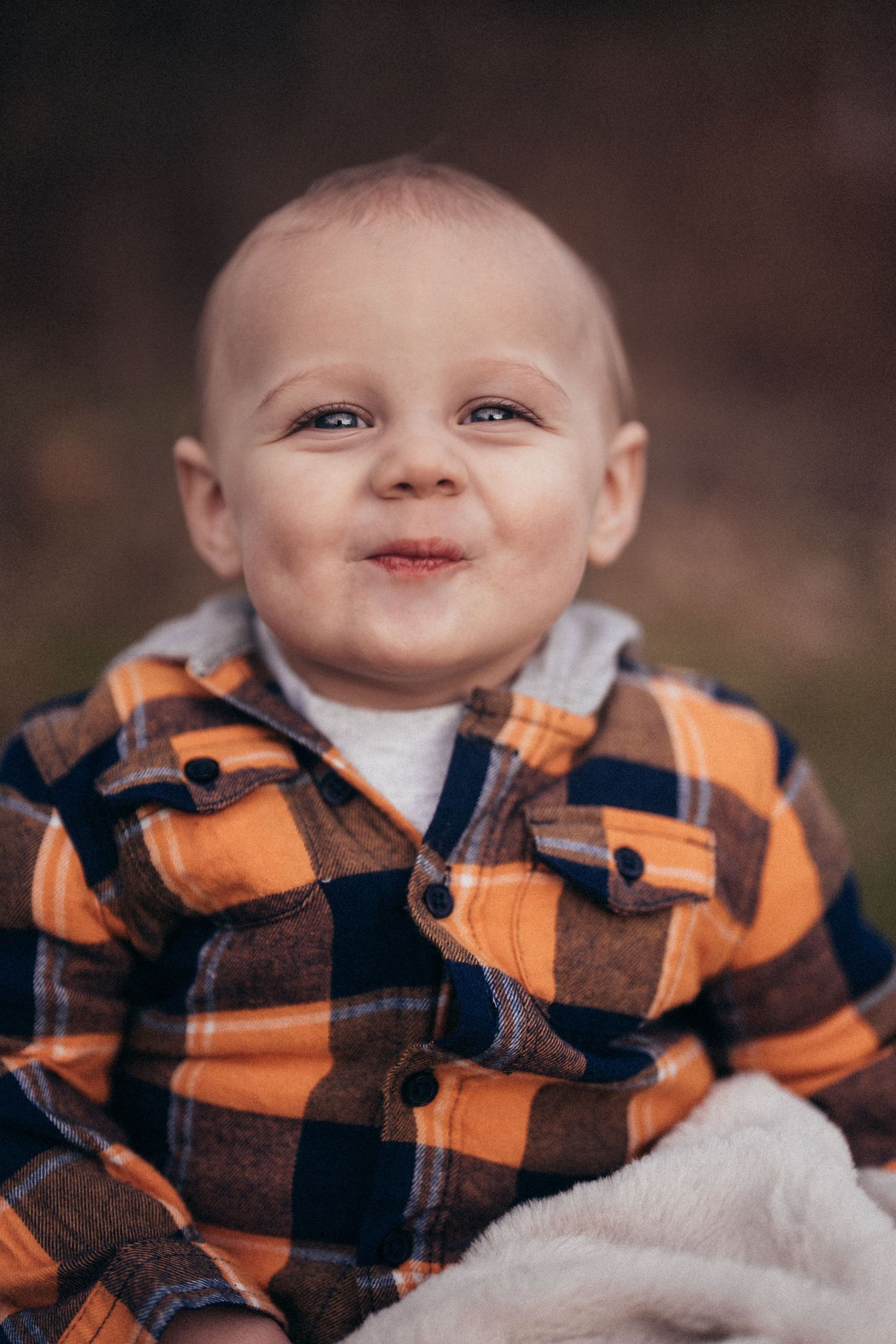 Smiling baby boy in orange plaid flannel shirt sitting outdoors on a soft blanket during fall season.