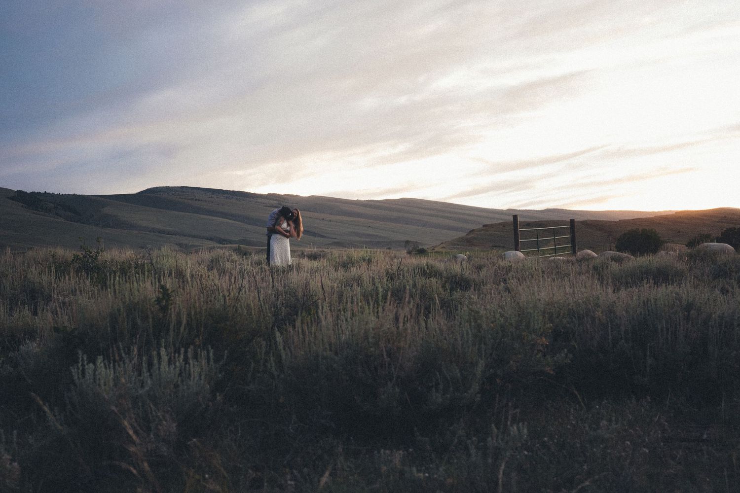 Couple embracing in a vast golden field at sunset with rolling hills and a wooden fence in the background.