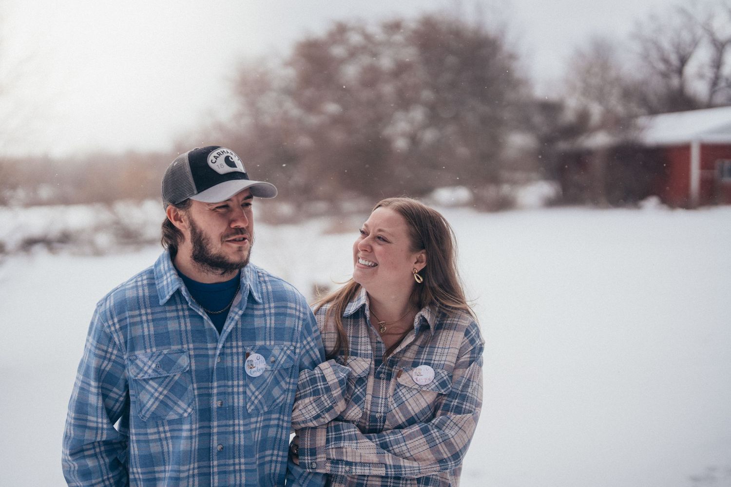 A couple in matching plaid shirts share a tender moment together in a snowy winter landscape.