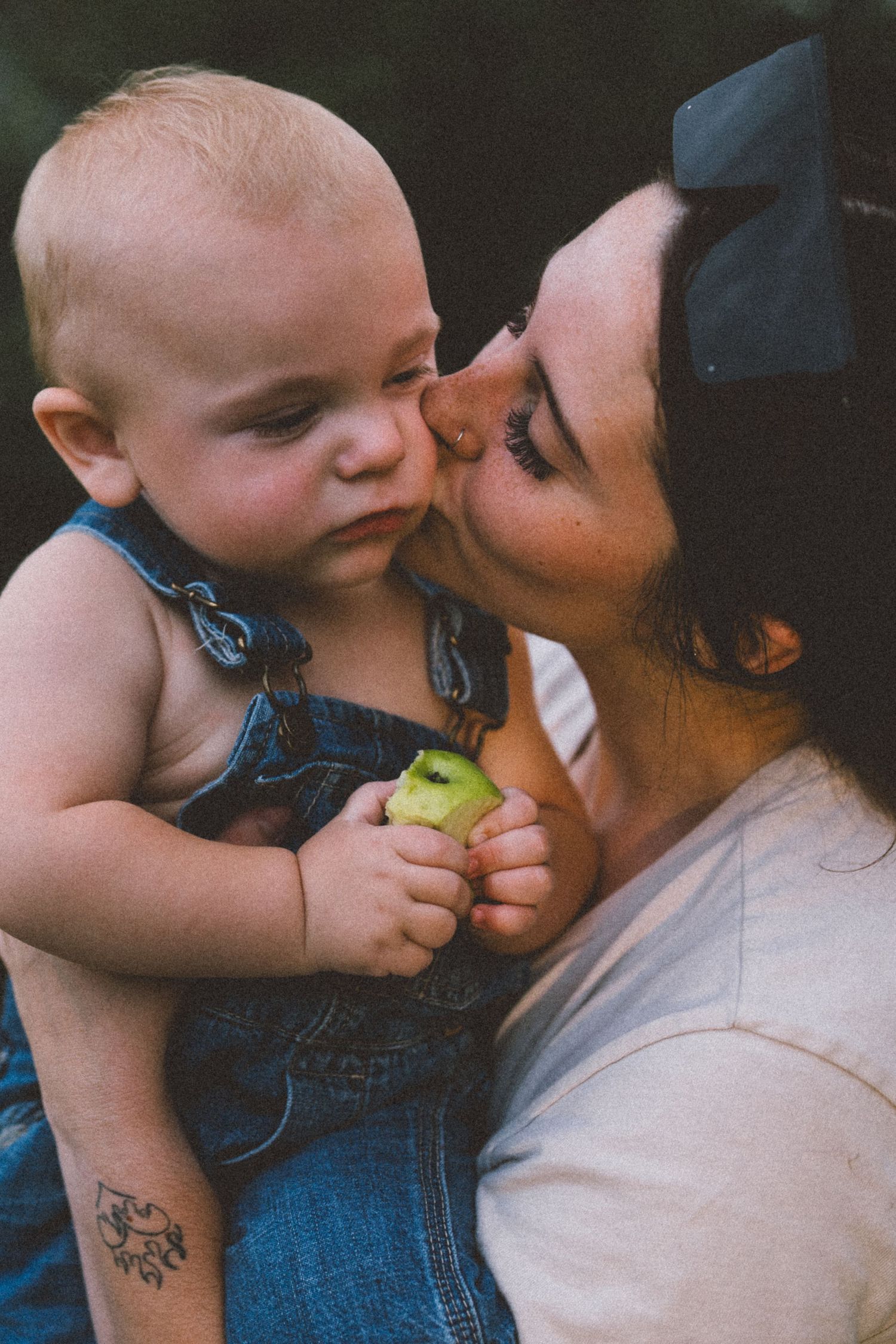 A tender moment between parent and baby in denim overalls holding a green apple.