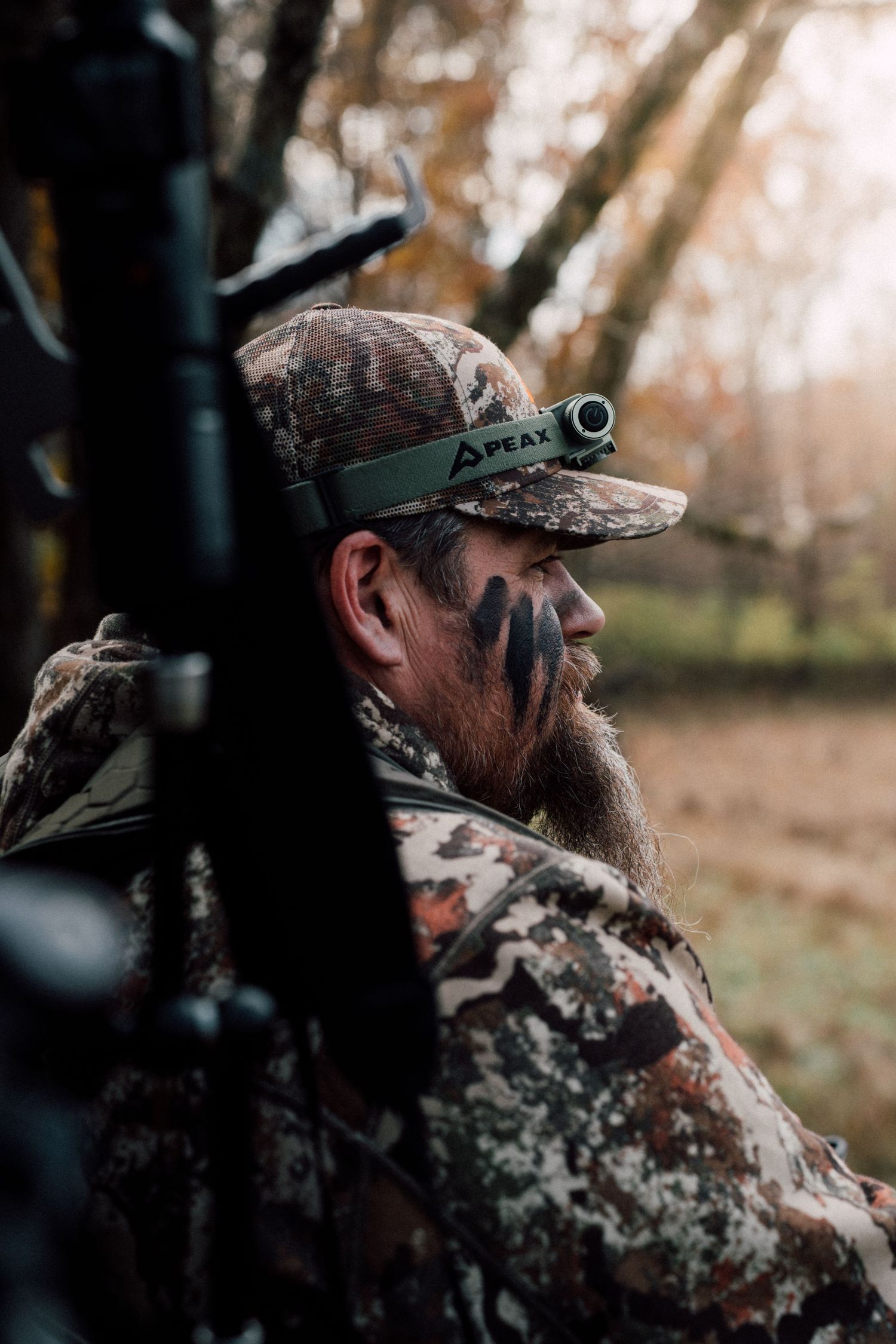 Bearded hunter in camouflage gear and PEAX headlamp sits quietly in autumn woods, face painted for concealment.