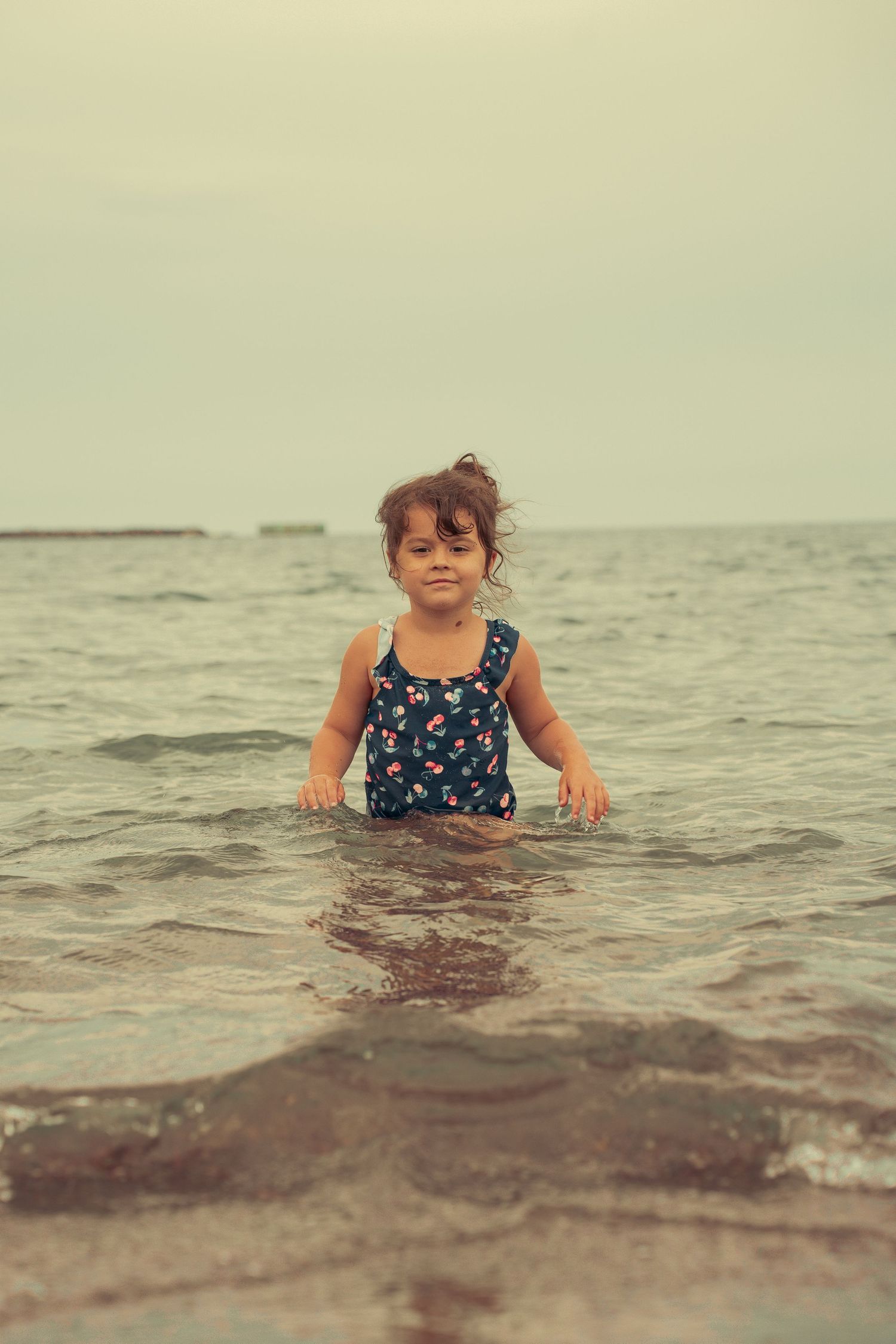 Young girl in floral swimsuit wading in ocean waves at beach, looking at camera with wet hair.