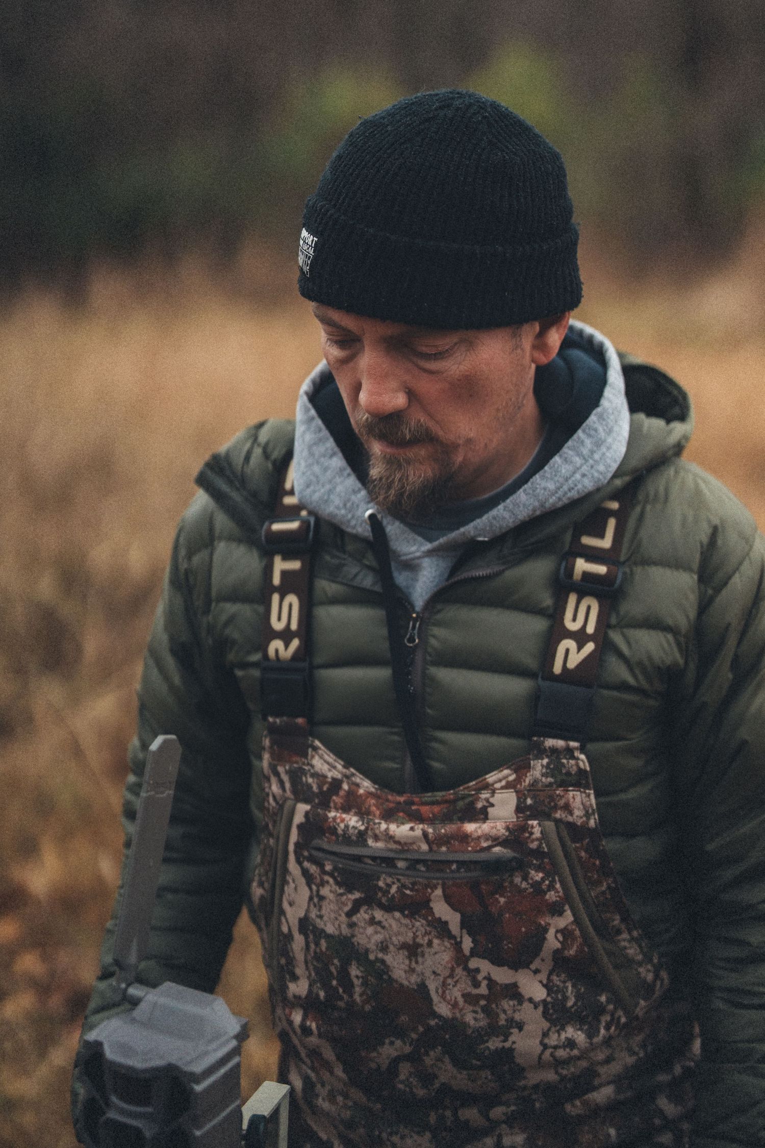 Man in camo overalls, green jacket, and black beanie holding binoculars outdoors in a field.
