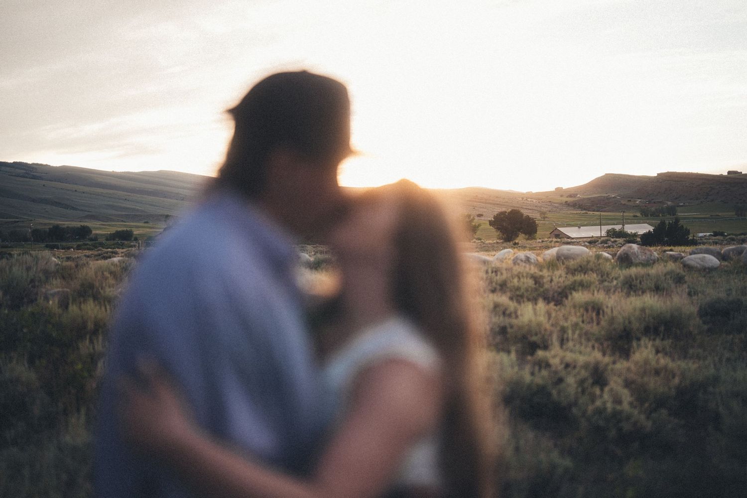 Couple sharing a romantic kiss at golden sunset in a scenic desert landscape with rolling hills and a lake in the background.