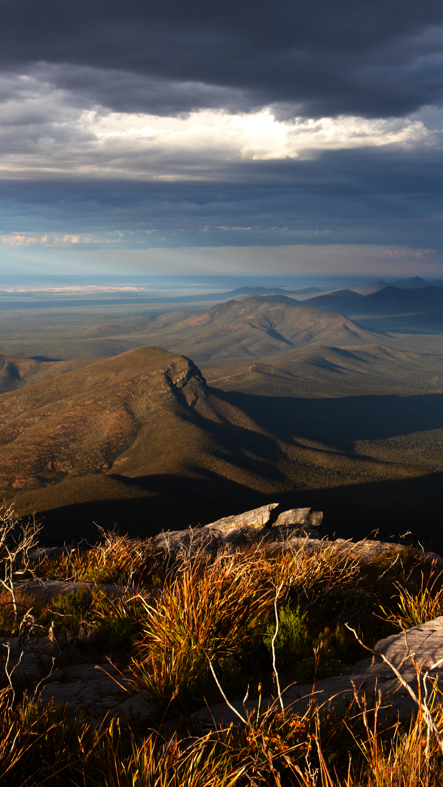 Sweeping mountain valley landscape with dramatic storm clouds and golden sunlight casting shadows over rugged terrain.