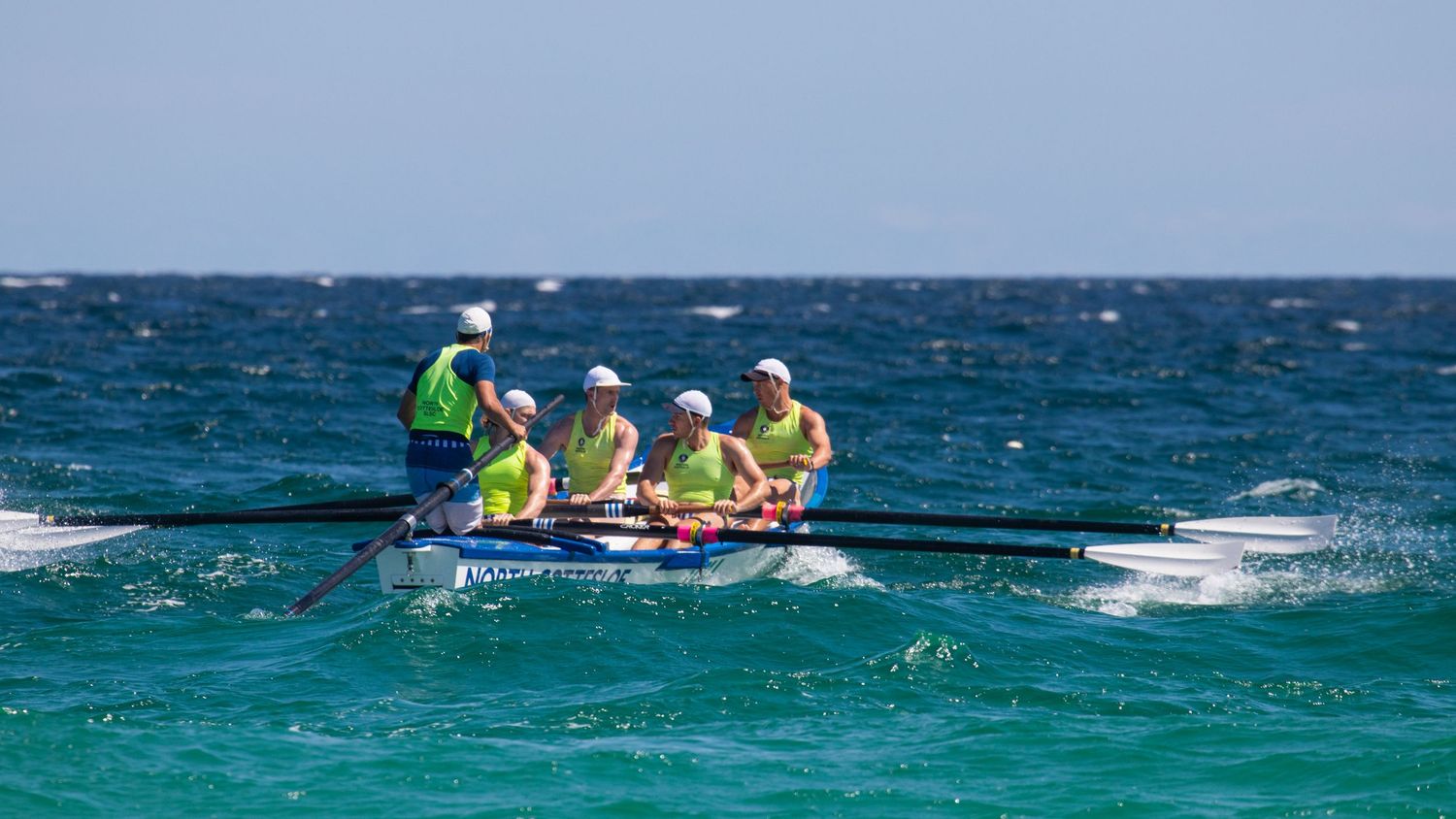 Four-person surf boat crew in yellow uniforms rowing through open ocean swells in a lifesaving race.