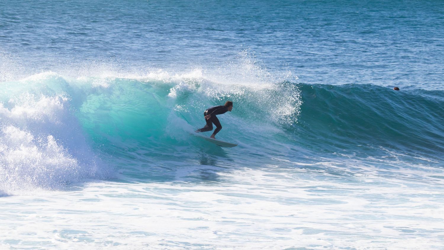 Surfer in black wetsuit carving through a beautiful turquoise barreling wave on a sunny clear day.