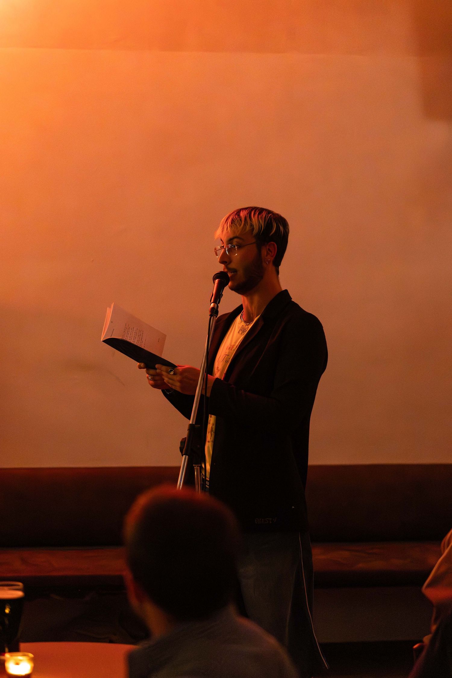 Bearded man in black jacket reads from paper at microphone during an indoor spoken word event.
