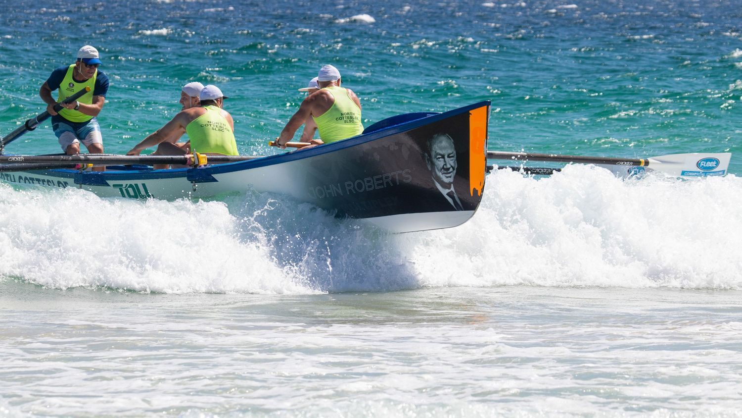 Surf boat crew rowing through breaking waves with portrait artwork displayed on the boat hull.