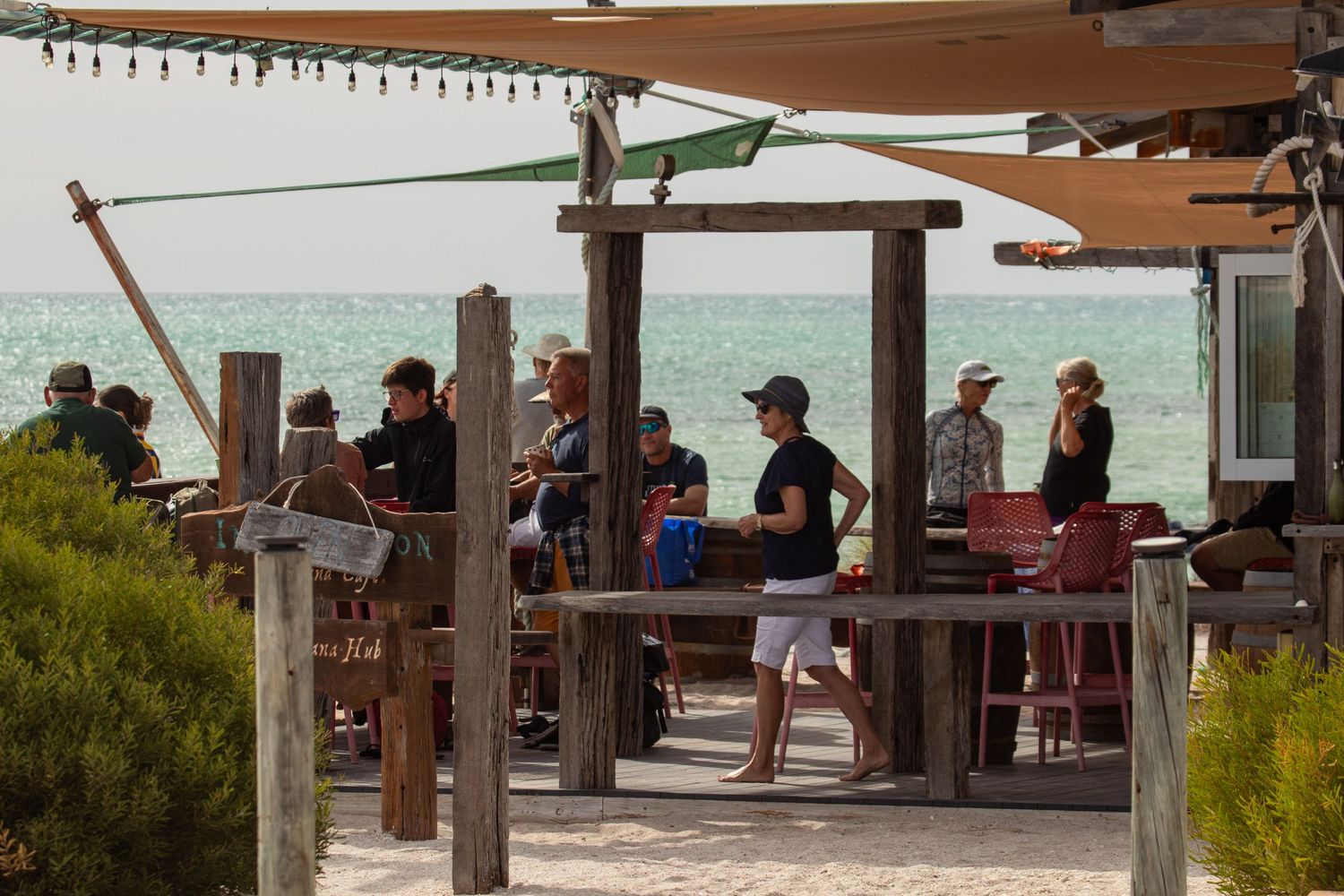 Groups of people socializing at a rustic wooden beachside bar under shade sails with turquoise water backdrop.