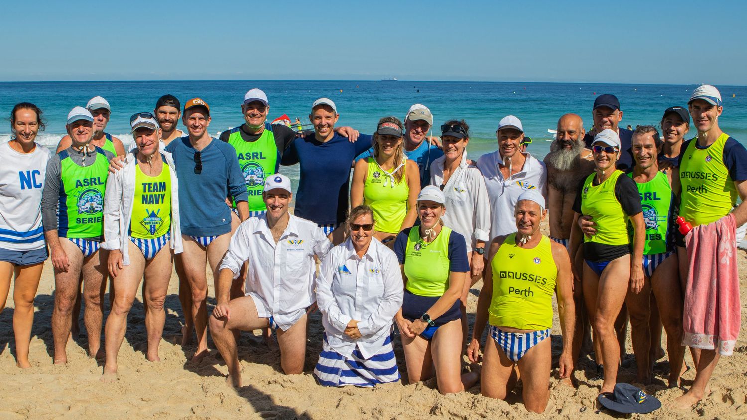 Large group of surf lifesaving competitors posing together on a sunny beach with ocean in the background.