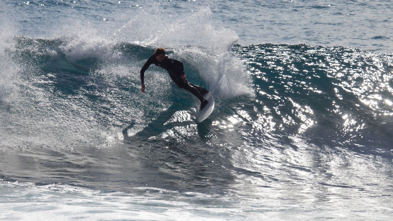 Surfer in black wetsuit carving a sharp turn on a white surfboard through a crashing ocean wave.