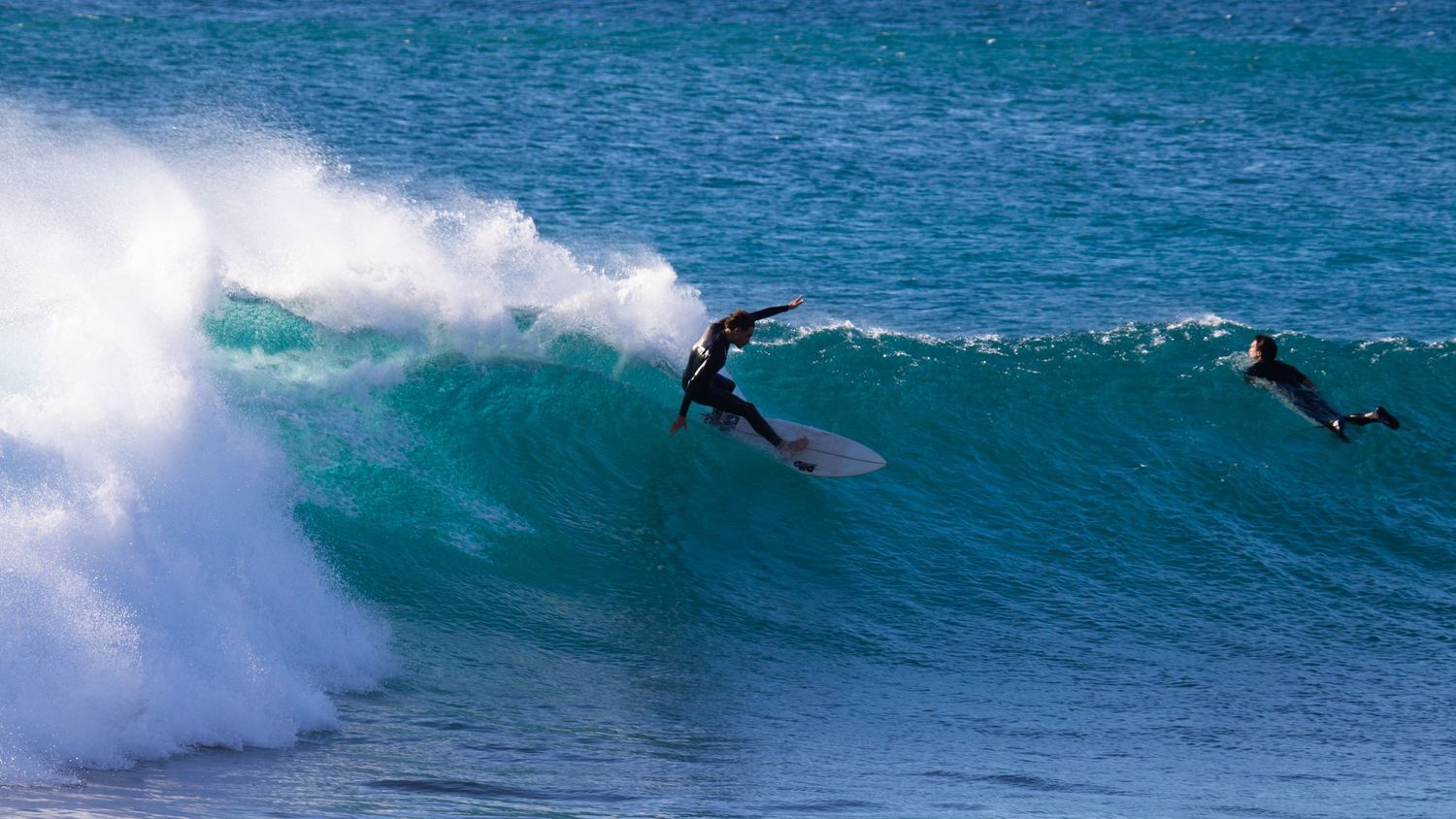 Two wetsuit surfers riding a large turquoise ocean wave.