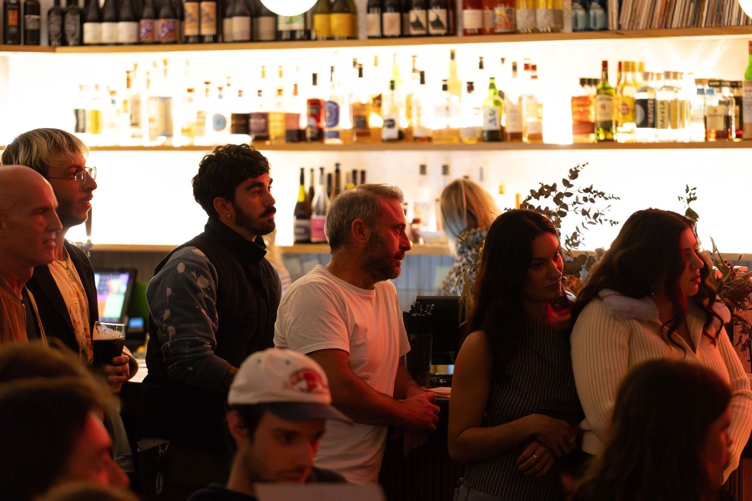 Crowd of people socializing at a trendy bar with shelves of bottles in the background.