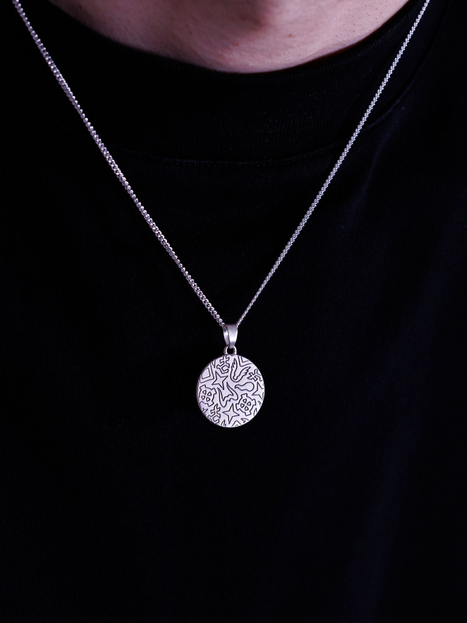Close-up of a silver round pendant necklace with detailed engraving worn on a black shirt.