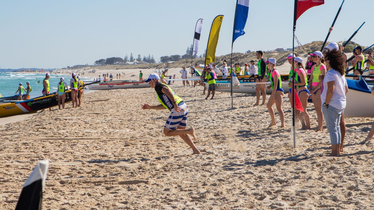 Surf lifesaving carnival participants running on beach with colorful flags and surf boats lined up.